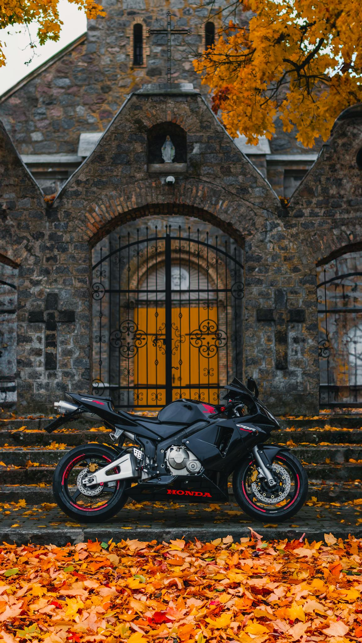 A black motorbike parked in front of a historic church gate amidst vibrant autumn leaves.