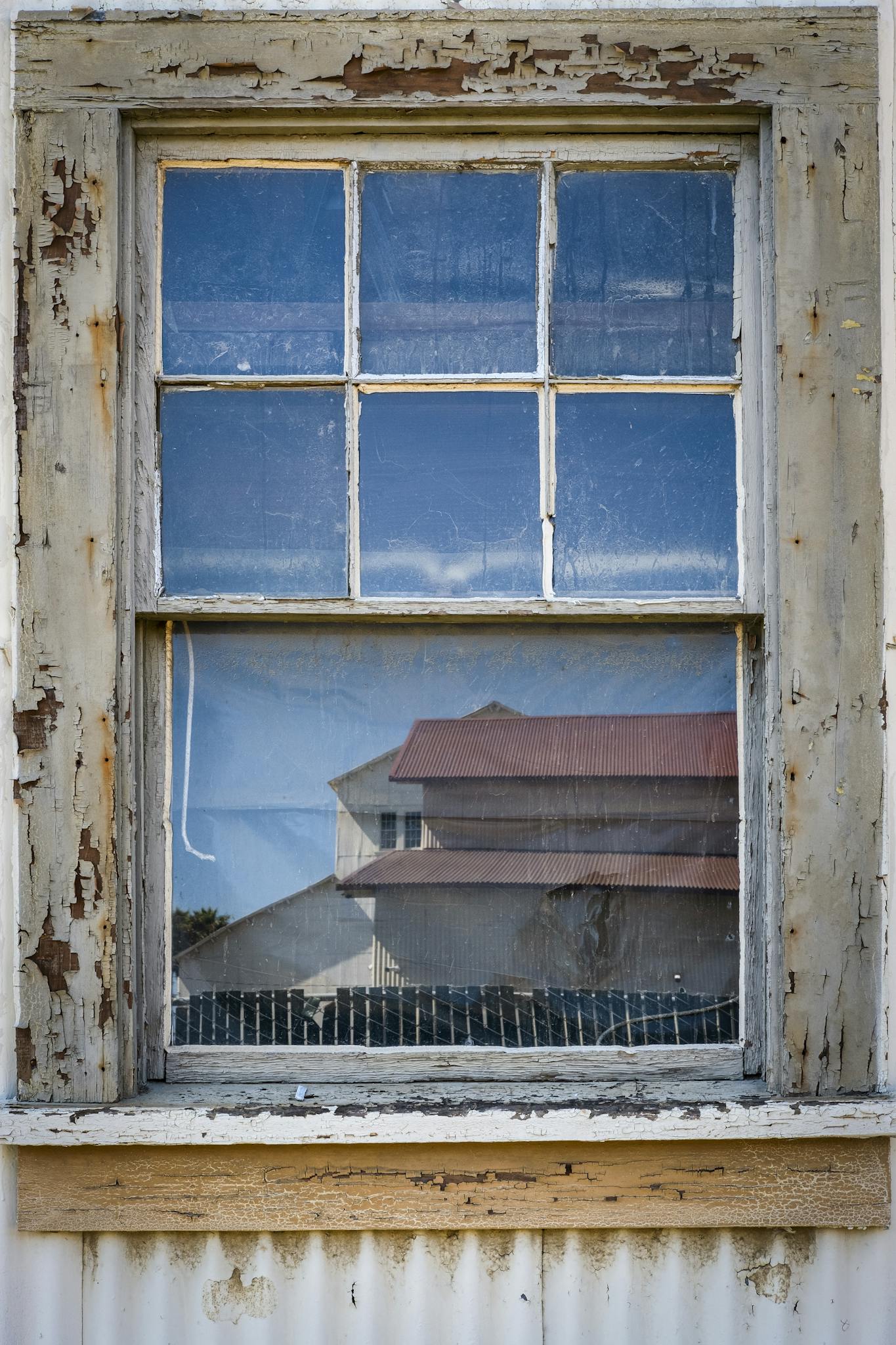 A close-up of an old wooden window reflecting a rural house with a red roof.