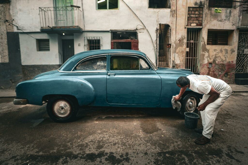 Man cleaning a classic blue car on a gritty urban street, depicting a vintage and rustic setting.