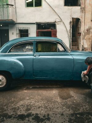 Man cleaning a classic blue car on a gritty urban street, depicting a vintage and rustic setting.